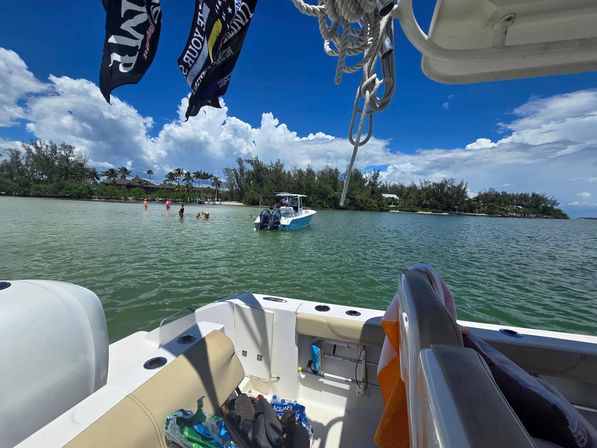 From a motorboat deck, view of a shallow turquoise bay with an anchored skiff and people wading near a palm-lined sandy shore under bright blue sky and puffy clouds, ropes and boat gear in the foreground.