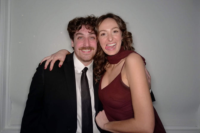 Playful portrait of two adults at a formal event — man in a black suit and tie smiling and woman in a maroon dress playfully sticking out her tongue with their arms around each other against a plain indoor backdrop.