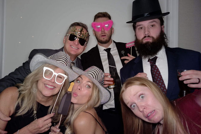 Six people in an indoor photo booth at a party wearing hats and playful props (paper glasses, crown, pink heart shades), holding drinks and making goofy faces
