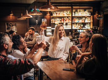 Friends raising pints and cheering in a cozy wood-lit pub, smiling around a table under pendant lights with a stocked bar in the background.