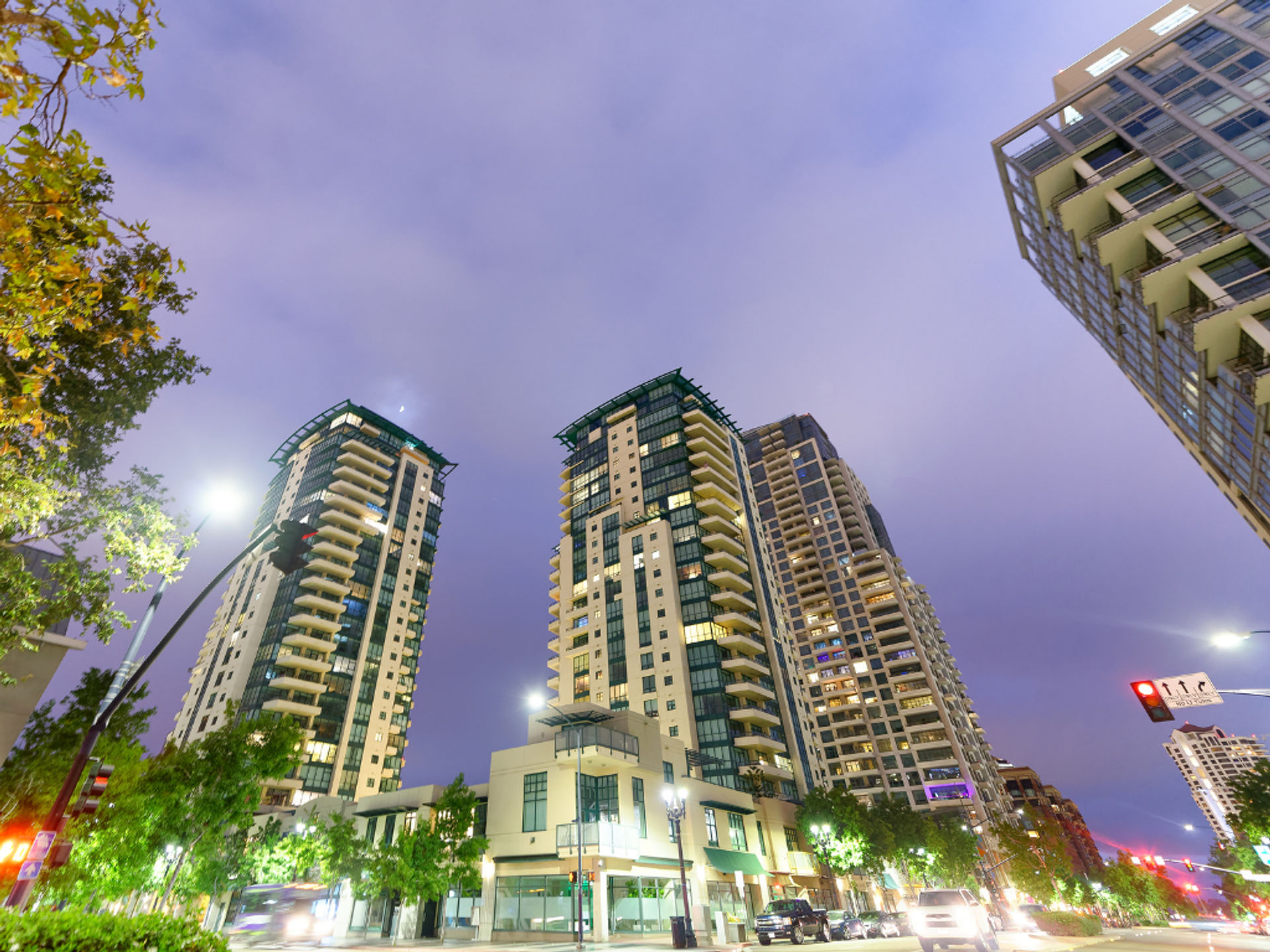 Upward view of modern downtown high-rise apartment towers glowing at dusk, with lit balconies, street-level shops, trees and cars on a busy city street under a purple evening sky.