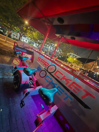 Nighttime outdoor pedal-powered bar with red canopy and neon lights, row of bicycle seats and pedals around a communal counter on a waterfront promenade with trees and illuminated bridge in the background.