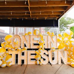 Giant illuminated marquee letters reading "ONE IN THE SUN" decorated with clusters of yellow-and-white beach balls on a covered urban patio event backdrop