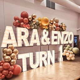Indoor party backdrop with oversized illuminated marquee letters and a festive balloon garland in rose gold, gold, peach and sage green announcing a first birthday.