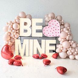 Valentine’s Day photo backdrop: illuminated “BE MINE” marquee letters with a pink polka-dot heart, clustered blush balloons on a white brick wall and scattered red heart-shaped foil balloons on the floor.