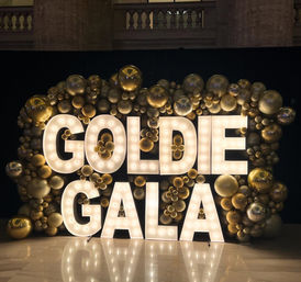 Illuminated marquee letters spelling GOLDIE GALA surrounded by clusters of metallic gold balloons of various sizes against a dark backdrop on a marble event floor, festive gala decor