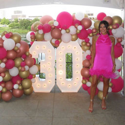 Person in a hot-pink mini dress posing beside illuminated marquee "30" surrounded by pink, gold and white balloon garlands at an outdoor riverside 30th birthday party.