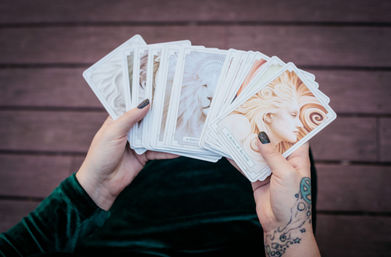 Hands with black glitter nails and a wrist tattoo fanning an illustrated tarot deck featuring ethereal female and lion portraits, photographed over a wooden deck.