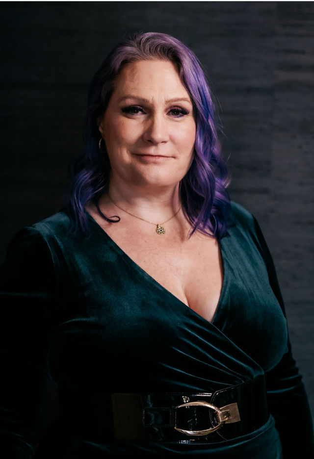 Studio headshot of a confident person with purple wavy hair, wearing a deep green velvet wrap dress, black patent waist belt and gold pendant necklace against a dark textured backdrop.