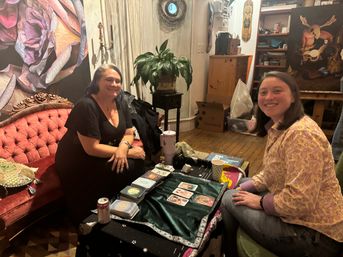 Two people smiling during a tarot card reading at a cozy bohemian art studio, with tarot cards on a velvet-covered table, a red tufted sofa, potted plant, wooden floors and colorful wall art.