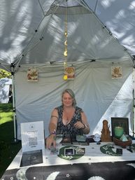 Person at an outdoor market tarot booth under a white canopy in a park, arranging tarot cards on a cloth-covered table with a hanging gold moon-phase mobile, sun and moon art prints, green candle, crystals and a wooden statue.