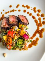 Grilled medium-rare steak slices atop a colorful summer bean-and-corn salad with cherry tomatoes, red onion, herbs and mustard vinaigrette drizzle on a white plate