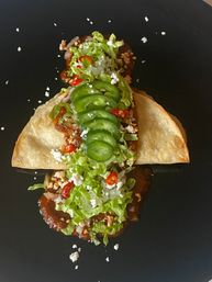 Mexican-style taco on a black plate: folded tortilla with spicy dark sauce, seasoned meat, shredded lettuce, thin jalapeño slices, red chili rings and crumbled white cheese.