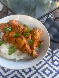Hearty orange chicken curry with drumsticks and carrots over steamed white rice, topped with cilantro, photographed on an outdoor patio with a glass table and patterned rug.