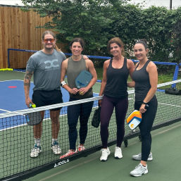 Four adults smiling and holding pickleball paddles on an outdoor fenced pickleball court at the net, ready to play amid trees and a wooden fence.