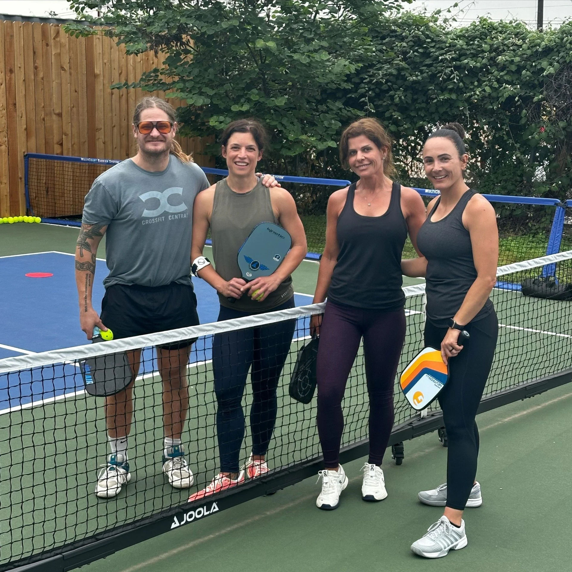 Four adults smiling and holding pickleball paddles on an outdoor fenced pickleball court at the net, ready to play amid trees and a wooden fence.
