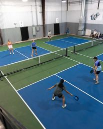 Overhead view of an indoor pickleball facility with multiple blue-and-green courts, players with paddles positioned for a doubles rally at the net in a recreational sports center.