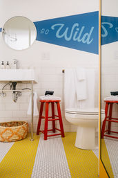Bright, playful bathroom with white subway tile, round mirror, blue 'Go Wild' wall graphic, red wooden stool, folded towels, woven basket and yellow-and-white penny-tile floor.
