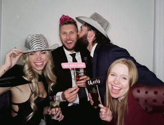 Four friends in a photo booth at a celebration, wearing sparkly hats and a crown, holding ‘We love you’ and ‘Cheers’ signs and cocktails, laughing and smiling.