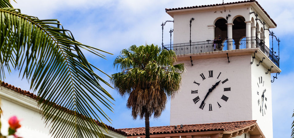 Mediterranean-style white clock tower with Roman numerals and arched observation balcony, terracotta roof and palm trees against a bright blue sky — sunny coastal vibe.