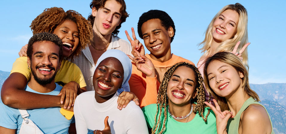Smiling diverse group of young friends posing outdoors with peace signs under a sunny blue sky and mountain backdrop