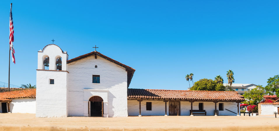 White adobe Spanish-style mission church with double bell tower, red tile roof, American flag and palm trees under a clear blue Southern California sky.