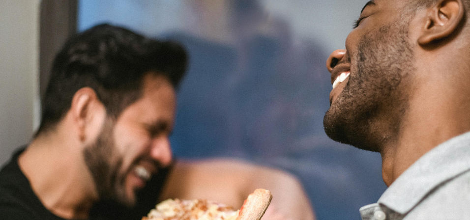 Close-up of two friends laughing and sharing a pizza slice in a cozy indoor setting