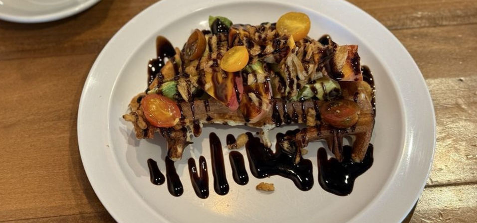 Brunch-ready savory waffle on white plate topped with sliced avocado, halved cherry tomatoes, shredded chicken and chopped walnuts, drizzled with dark balsamic glaze on a wooden table