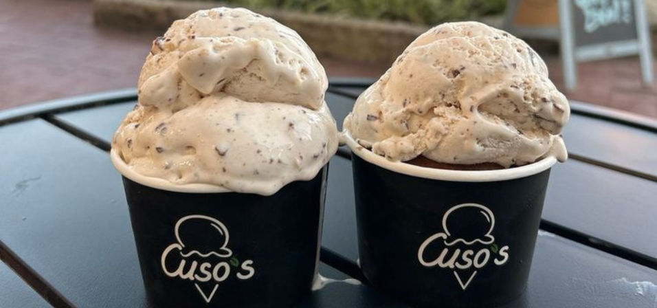 Two overflowing scoops of creamy vanilla ice cream with chocolate chunks in black paper cups on an outdoor café table