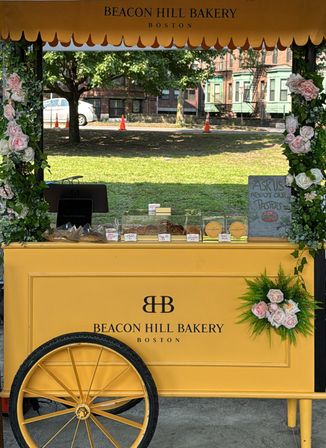 Bright yellow vintage-style bakery cart with rose garlands, pastries in clear cases, and a chalk sign at an outdoor Boston park market.