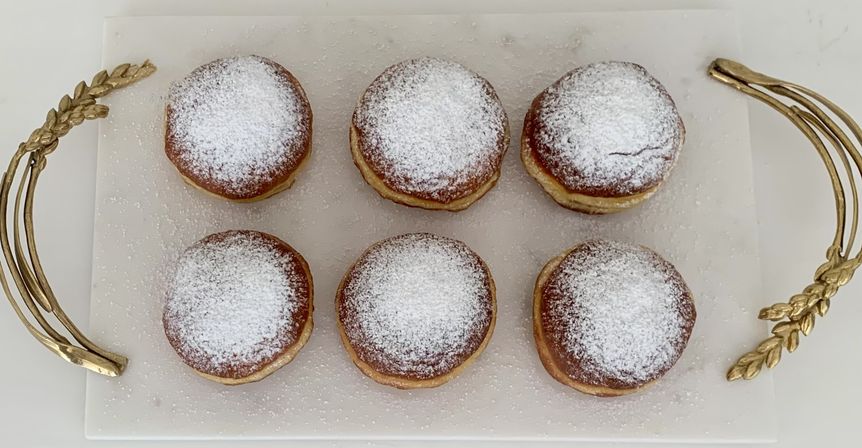 Top-down view of six round powdered-sugar donuts arranged in two neat rows on a white serving board with decorative gold handles.