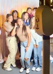 Seven friends posing at an indoor photo booth with a white drape backdrop, making peace signs and playful poses in casual jeans and sneakers.