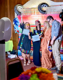 Indoor birthday party photo booth with four adults posing in front of a colorful striped backdrop, wearing leis and sunglasses and holding “PHOTO BOMB” and “BEST BIRTHDAY PARTY EVER” signs with festive balloons and fringe decor.