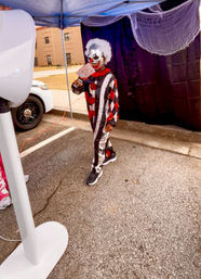 Person in a scary clown costume with white wig and red-and-white face paint holding a fake bloody cleaver under a canopy at an outdoor Halloween setup in a parking lot with a dark backdrop and nearby car.