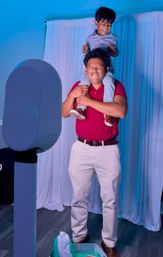 Adult in a red polo and beige pants grinning with eyes closed while carrying a laughing child on his shoulders in front of a photo booth, blue-purple party lighting and white draped backdrop.