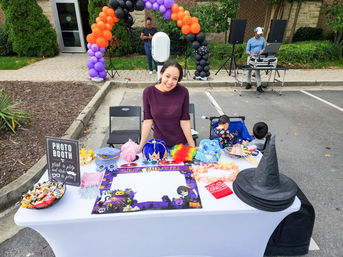 Outdoor Halloween photo booth setup in a parking lot with props (witch hats, colorful wigs, masks, candy), a smiling attendant at the table, purple-orange balloon arch and DJ in the background.