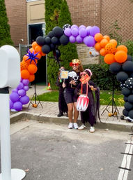 Group of three people posing at an outdoor Halloween photo booth under a purple, orange and black balloon arch by a brick building, wearing playful crowns and sunglasses and holding festive props in a parking-lot area