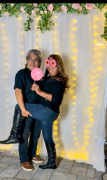 Couple posing in a party photo booth in front of a white curtain backdrop with hanging fairy lights and pink rose garland; woman wearing sunflower novelty glasses and black boots lifts her leg while man holds a pink party sign.