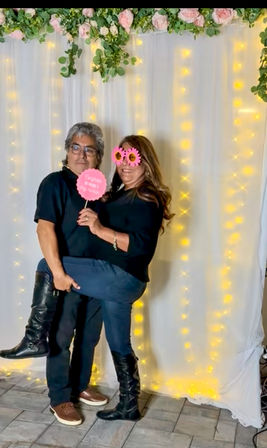 Couple posing in a party photo booth in front of a white curtain backdrop with hanging fairy lights and pink rose garland; woman wearing sunflower novelty glasses and black boots lifts her leg while man holds a pink party sign.