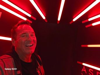 Smiling man taking a selfie inside a red neon light installation with glowing horizontal tubes radiating from walls and ceiling.
