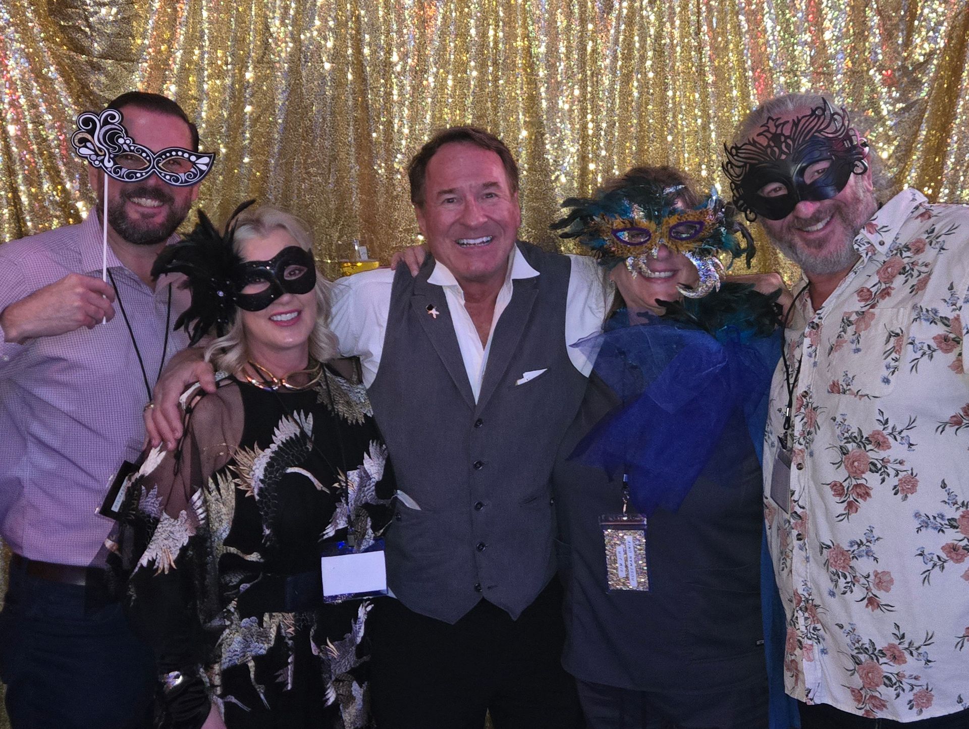 Five adults in festive attire wearing colorful masquerade masks and holding props, smiling together in a group photo in front of a gold sequin backdrop at an indoor party.
