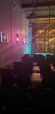 Cozy industrial loft event space with rows of chairs and small tables, pink and teal stage lights on an exposed brick wall, factory-style windows and hanging pendant lights.