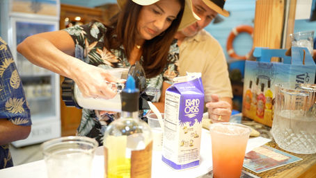 Two people pouring a frothy tropical cocktail at a beach-style bar counter with a mixer carton, liquor bottles and a pink iced drink — lively island vacation vibe