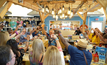 Lively distillery tasting room with wooden beams and hanging lights, a group of adults gathered around a bar raising shot glasses for a celebratory moonshine tasting.