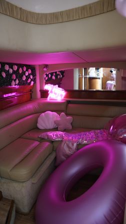 Pink-lit yacht cabin lounge with curved tan leather seating, floral wall panel, neon pink sign, shell and star-shaped pillows, sequin throw and large inflatable pink tube.