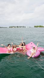 Five friends in pink bikinis laughing on a giant pink inflatable and donut tube in a coastal bay with boats and palm-lined shoreline — summer boating scene.
