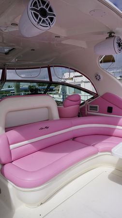 Curved pink-and-white yacht interior with plush banquette seating, pink captain’s chair, overhead marine speakers and marina visible through wraparound windows
