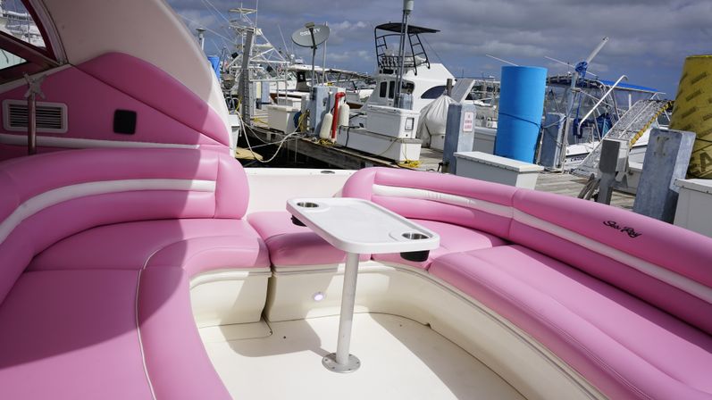 Pink curved vinyl seating with a small white table and cup holders on a boat, docked at a busy marina with fishing boats and docks under a cloudy sky.