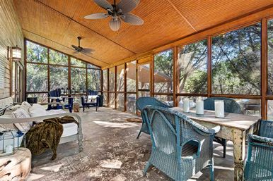 Sunlit rustic screened porch with beadboard wood ceiling and ceiling fans, shabby-chic dining table with blue wicker chairs, cozy daybed and lounge seating overlooking a wooded backyard
