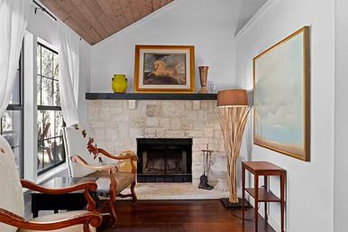 Cozy living room with stone fireplace and black mantel, vaulted wood ceiling, large mullioned window with white curtains, carved wooden armchairs on dark hardwood floors, tall woven floor lamp and framed wall art.